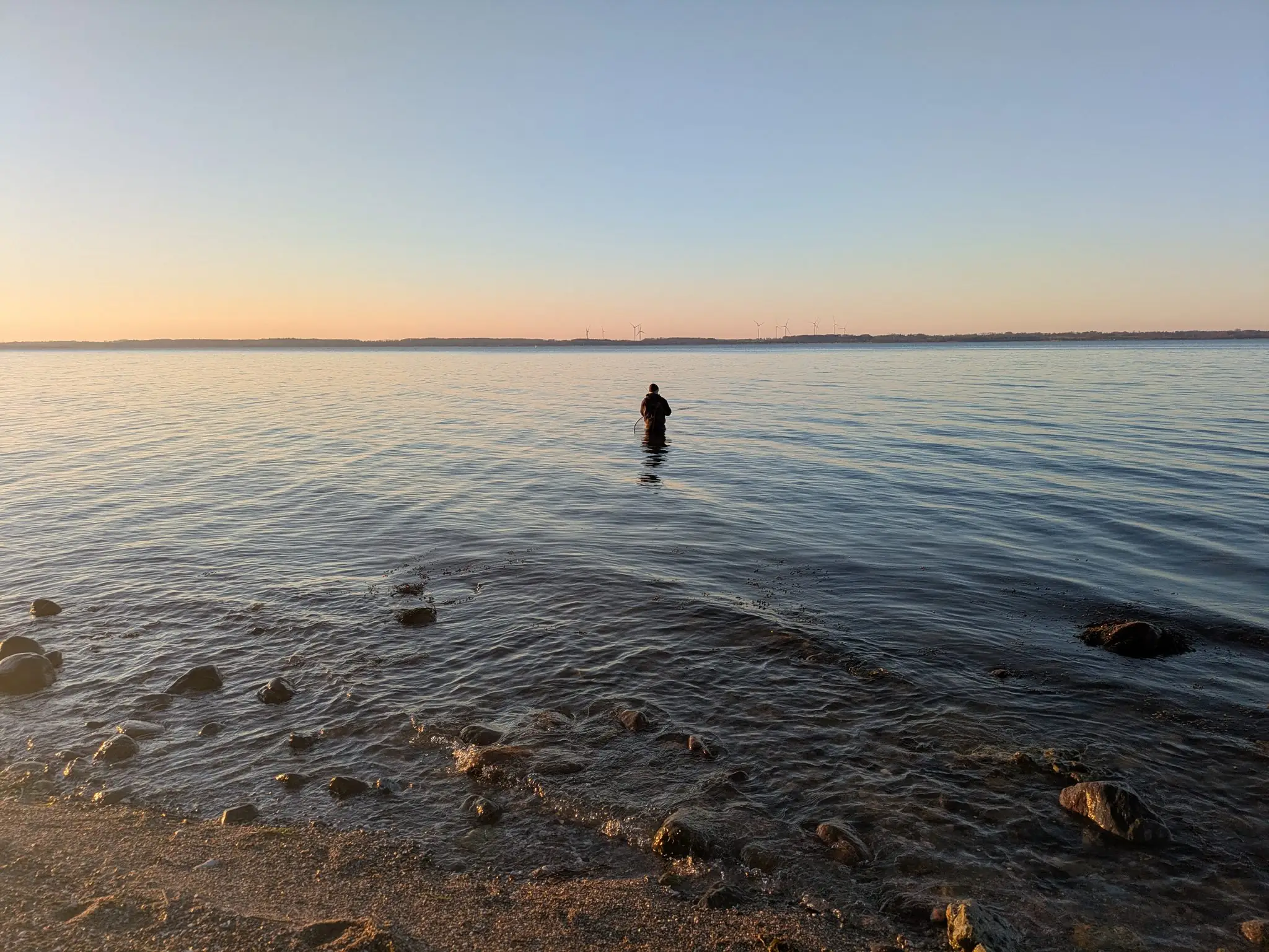 Angler steht im flachen Wasser des Wittensees beim Angeln, Sonnenuntergang, Windräder am Horizont