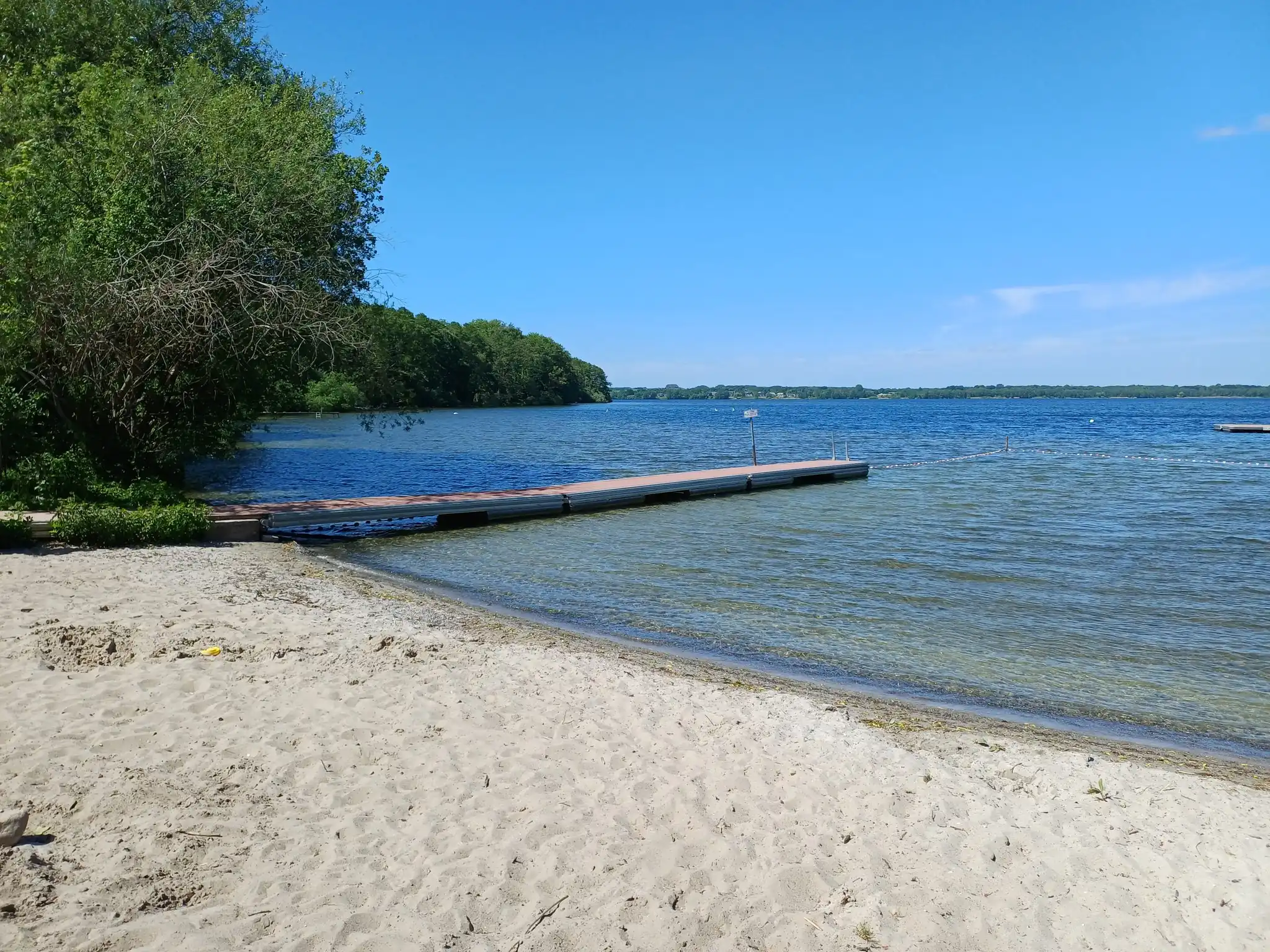 Sandstrand am Wittensee mit Badesteg und klarem Wasser, im Hintergrund bewaldete Uferböschung