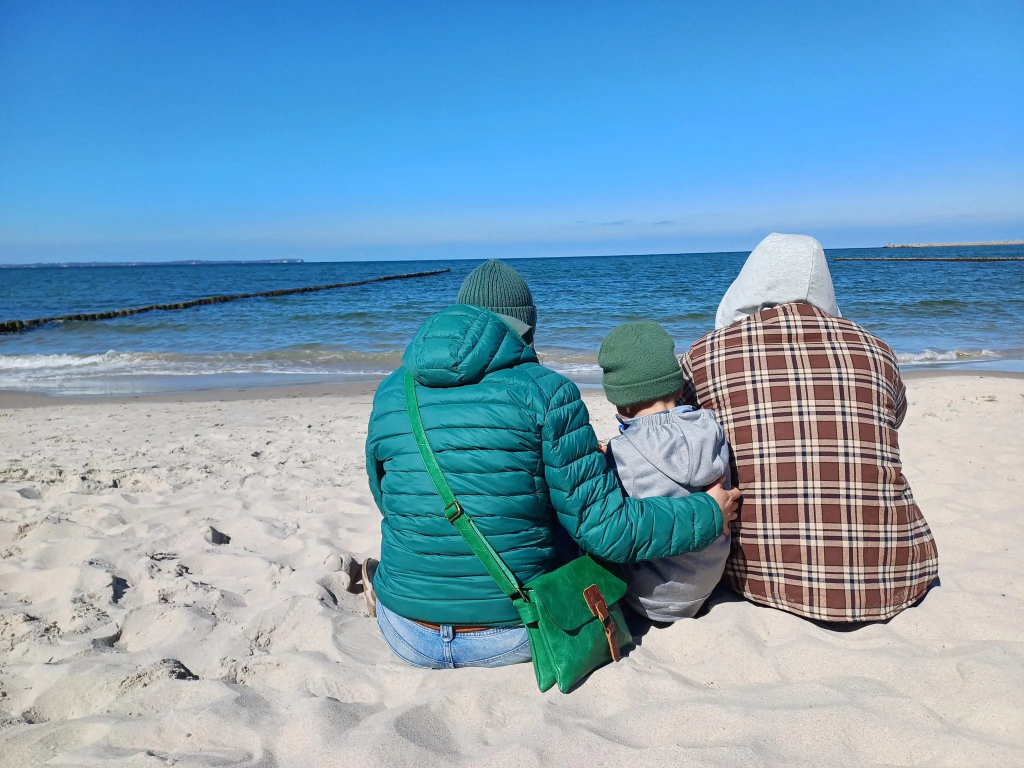 Familie sitzt am Sandstrand der Ostsee und blickt aufs Meer, Buhnen im Hintergrund