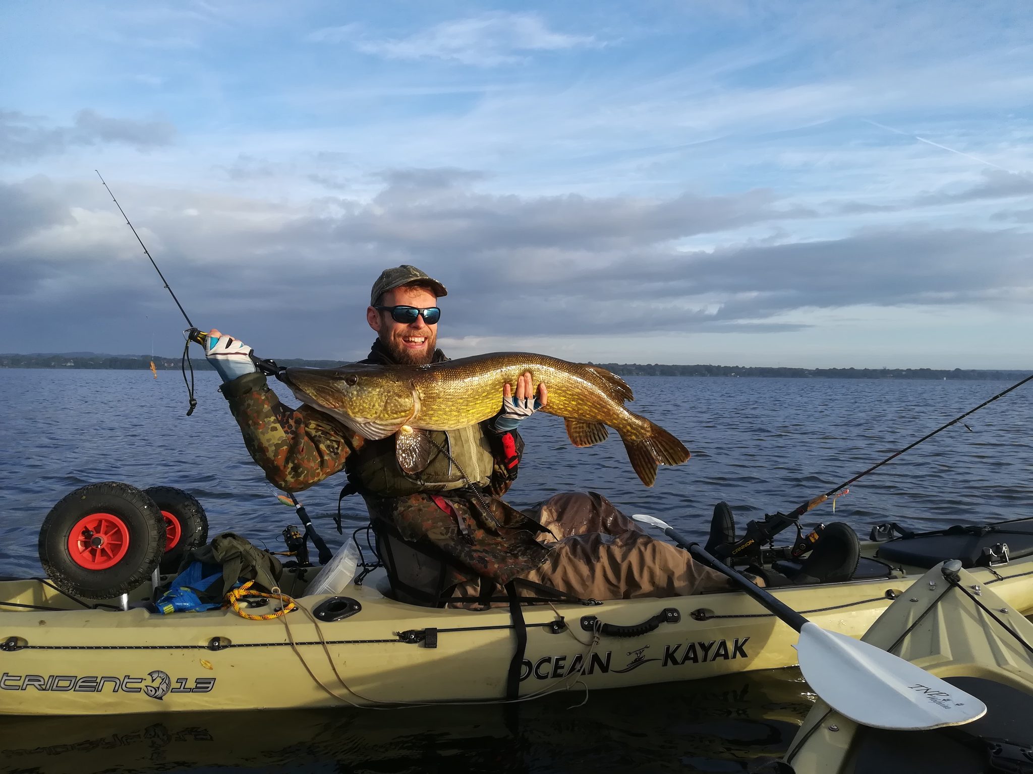 Angler im Kajak hält einen großen Hecht in die Kamera, im Hintergrund ruhiges Seewasser und bewölkter Himmel.