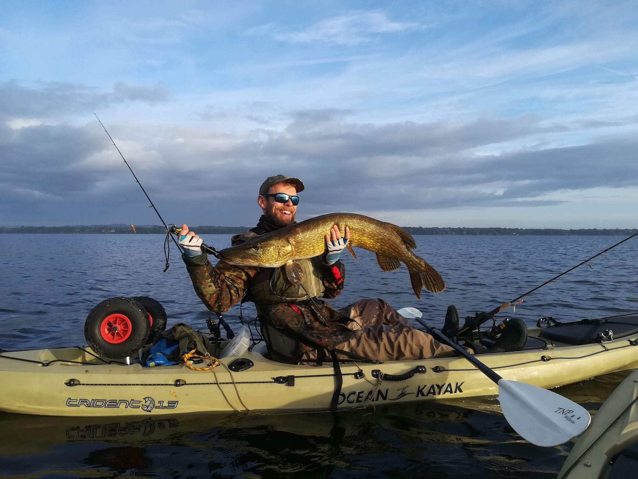 Angler im Kajak hält einen großen Hecht in die Kamera, im Hintergrund ruhiges Seewasser und Wolkenhimmel.