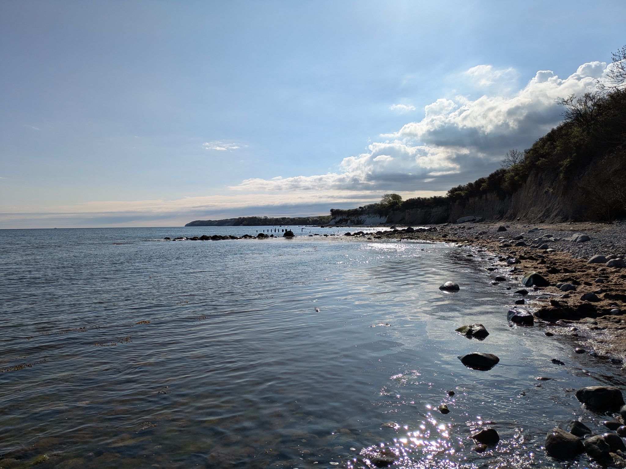 Steiniger Ostseestrand mit Steilküste und bewölktem Himmel, ruhiges Wasser im Sonnenlicht