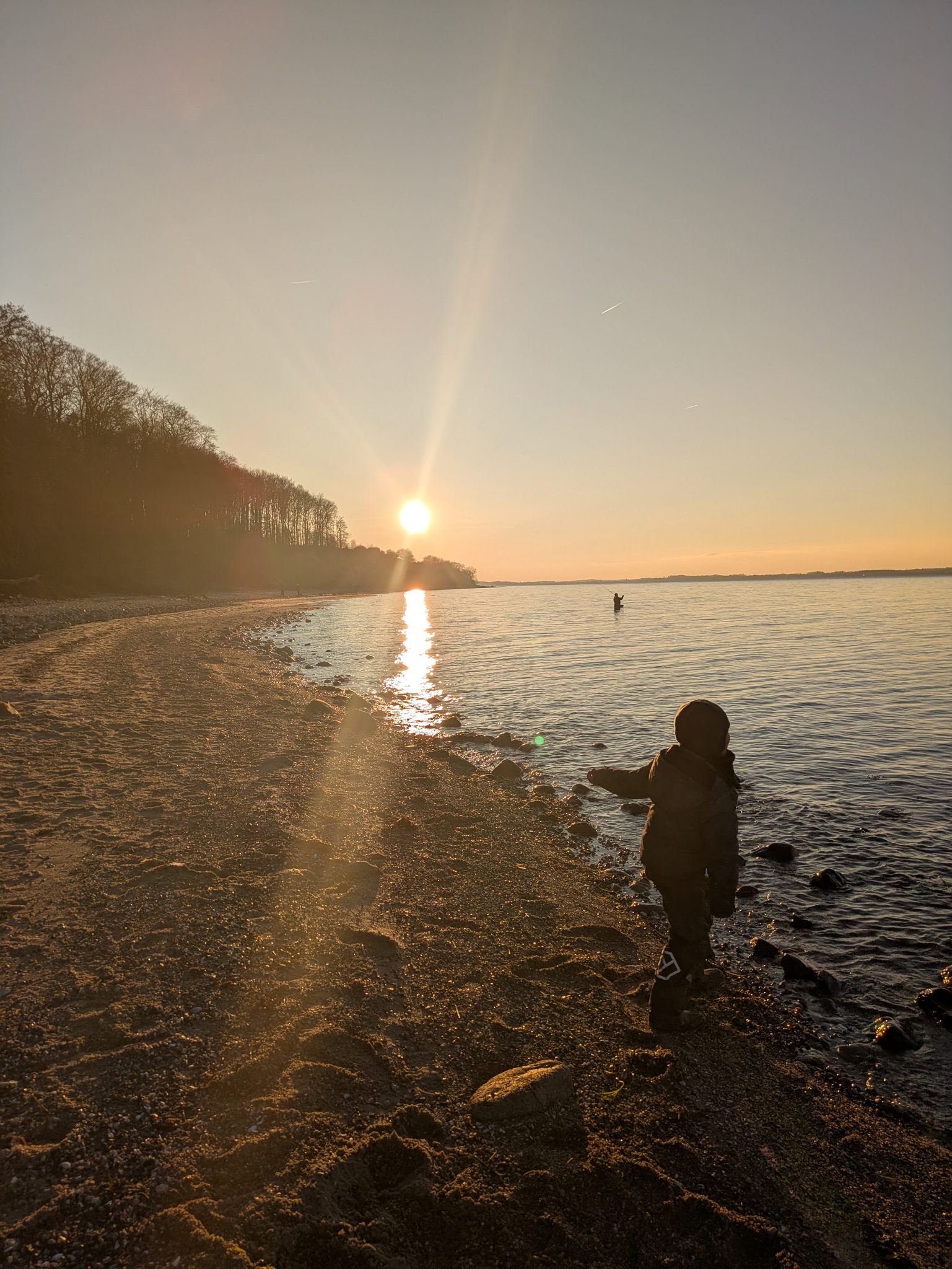 Kind in Winterjacke steht am Seeufer des Wittensees im Sonnenuntergang, im Hintergrund ein Angler im Wasser