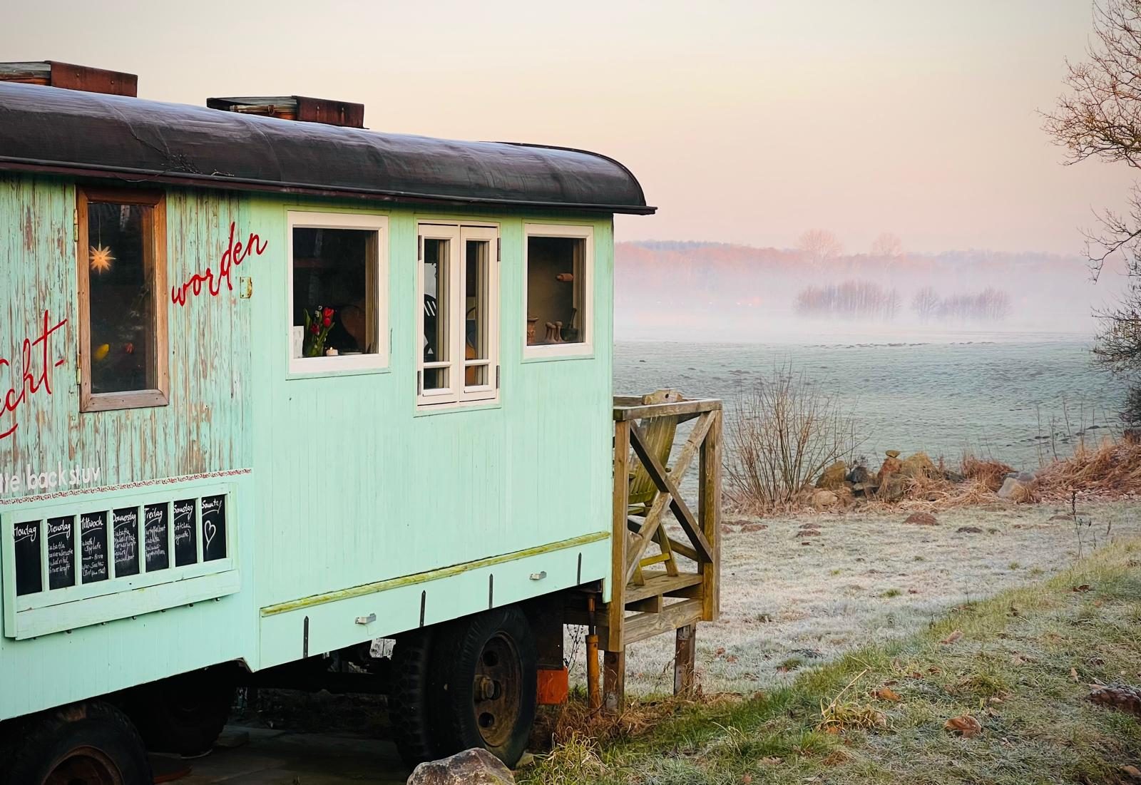 Mintgrüner historischer Bauwagen mit Holztreppe im Morgennebel, Wittensee im Hintergrund