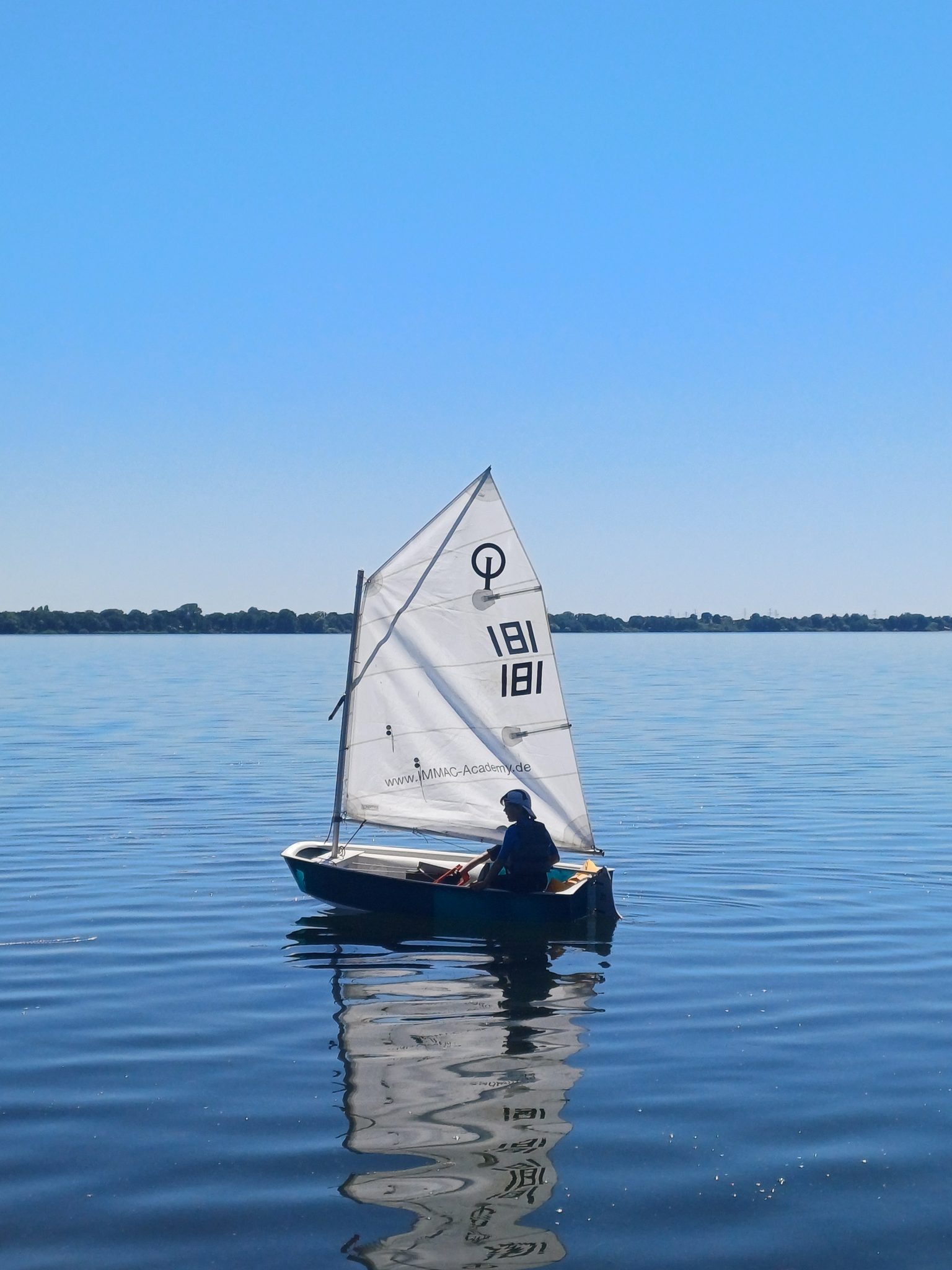 Kind segelt auf einem Optimisten-Dingi bei Sonnenschein auf dem Wittensee, spiegelglattes Wasser