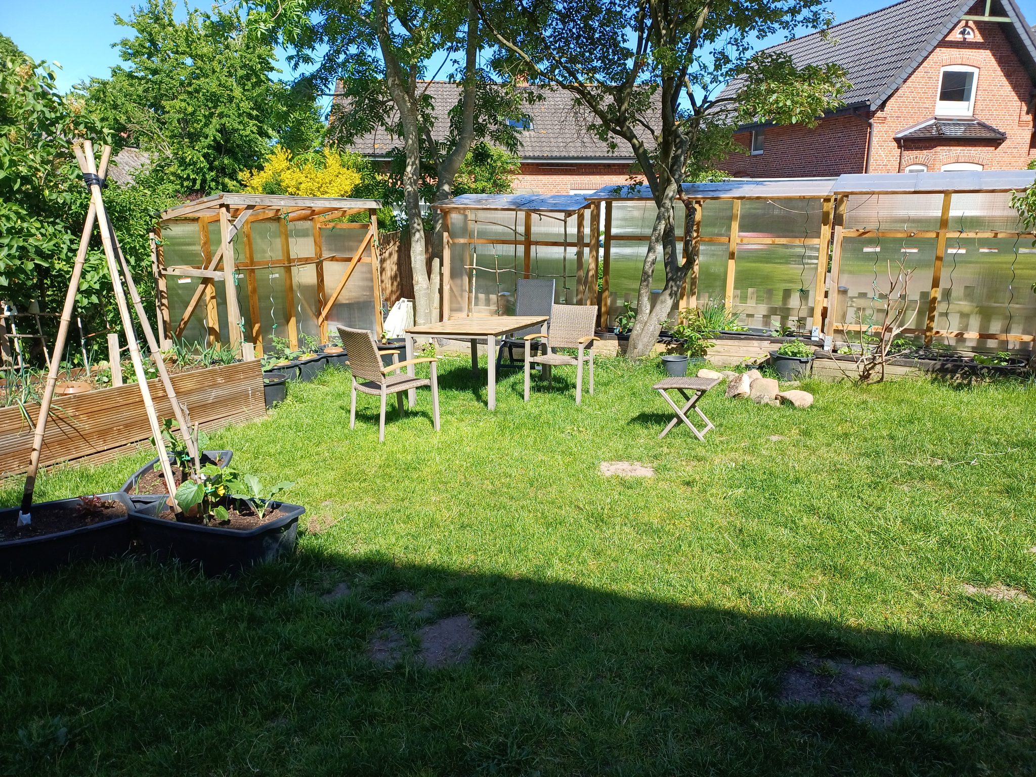 Garten mit Gewächshaus, Hochbeet, Gartengarnitur und grünem Rasen vor rotem Backsteinhaus