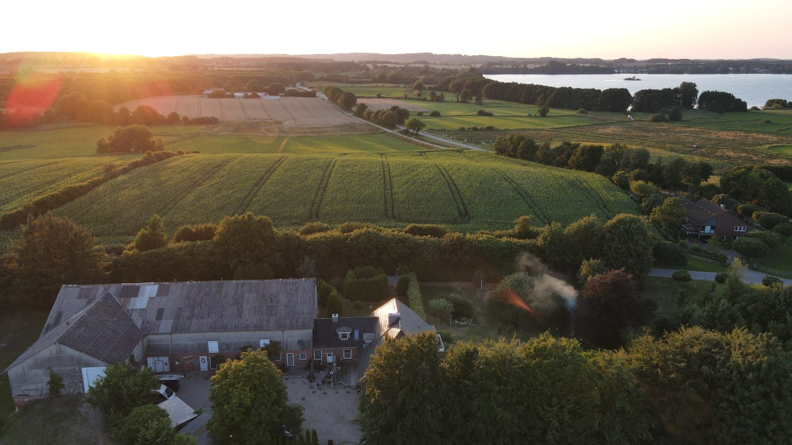 Luftaufnahme von Hof Gayenberg im Sonnenuntergang, umgeben von Feldern und Bäumen, Wittensee im Hintergrund