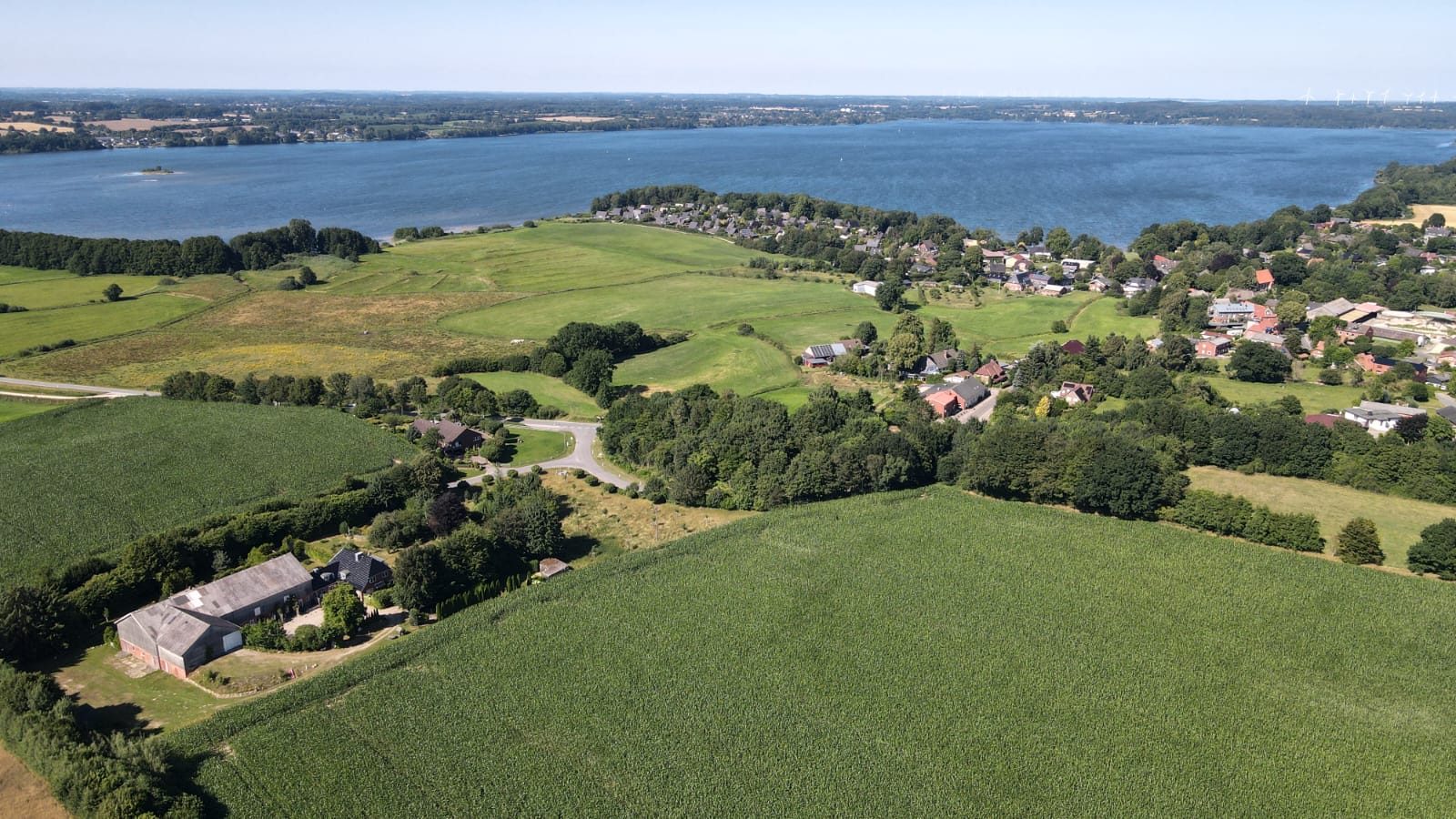 Luftaufnahme von Hof Gayenberg mit Feldern, umliegender Ortschaft und dem Wittensee im Hintergrund