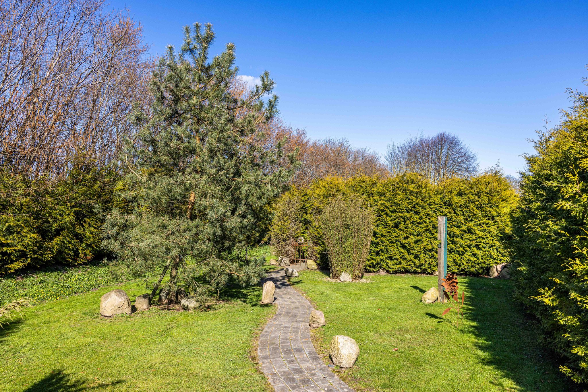 Gepflasterter Gartenweg mit Findlingen, Kiefern, Thuja-Hecken und Metallskulpturen unter blauem Himmel