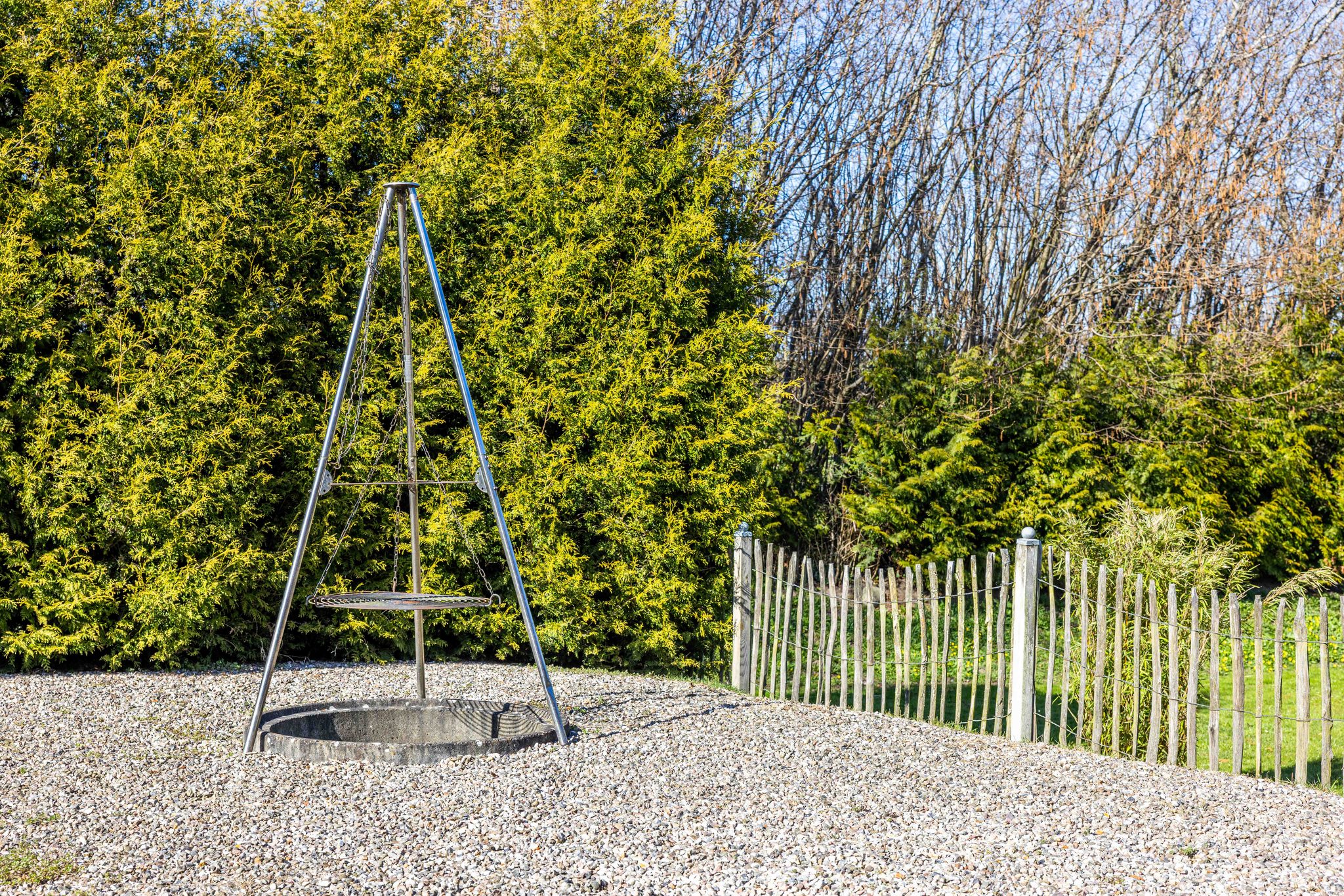 Dreibein-Schwenkgrill aus Edelstahl auf Kiesfläche, dahinter Thujenhecke und Holzzaun im Garten