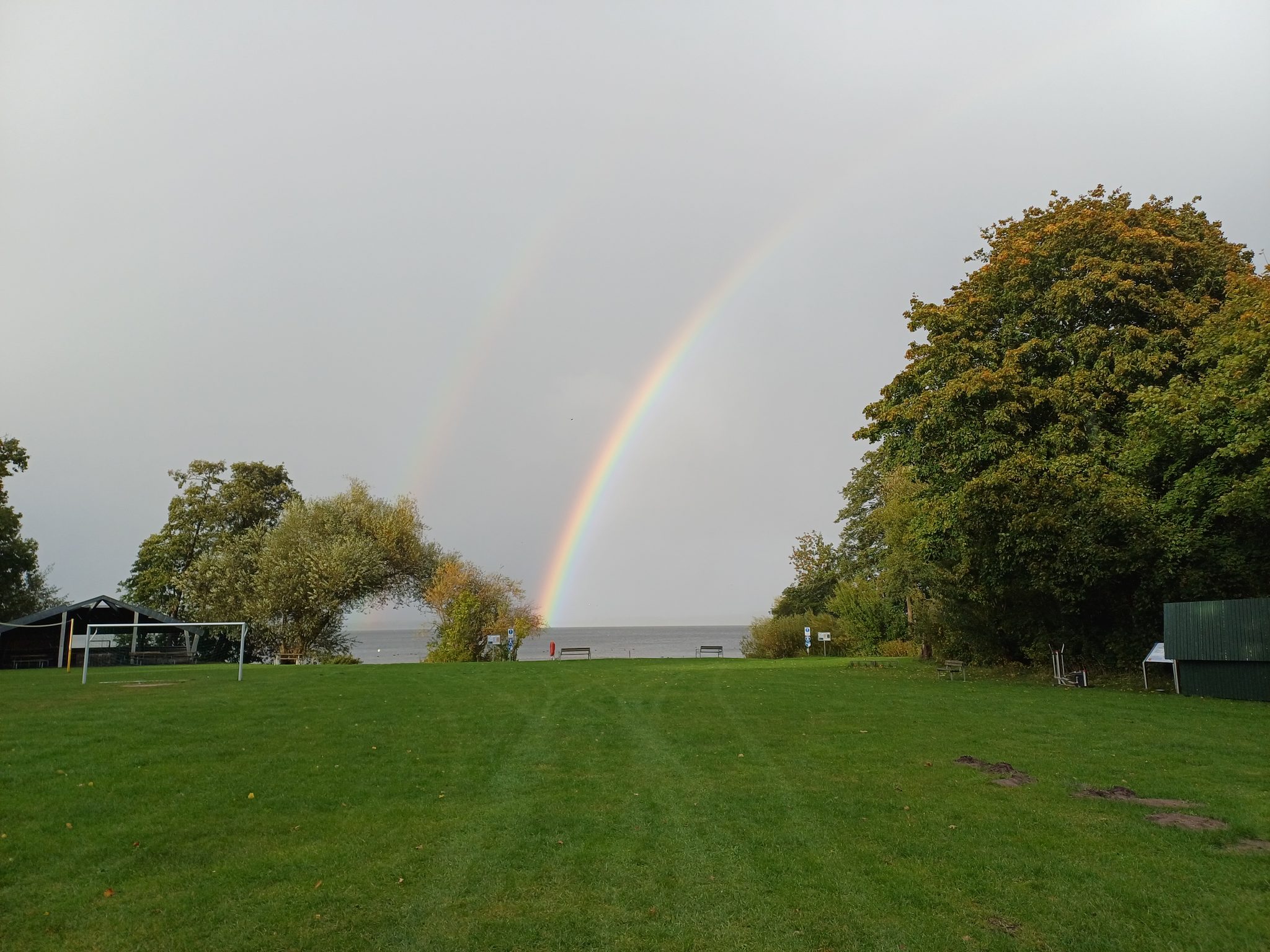 Doppelter Regenbogen über dem Wittensee, grüne Wiese mit Fußballtor und herbstlichen Bäumen im Vordergrund