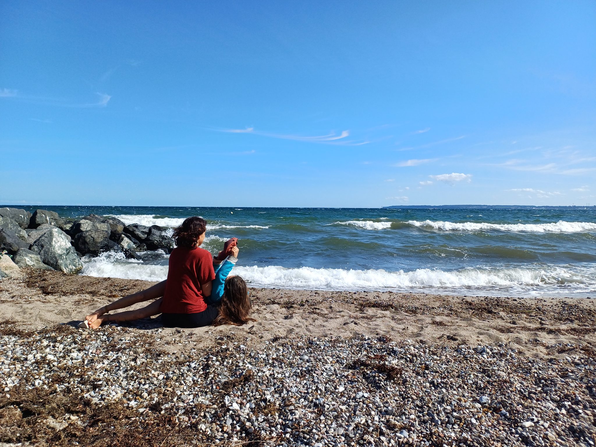 Frau und Kind sitzen am Kiesstrand der Ostsee und schauen auf die Wellen