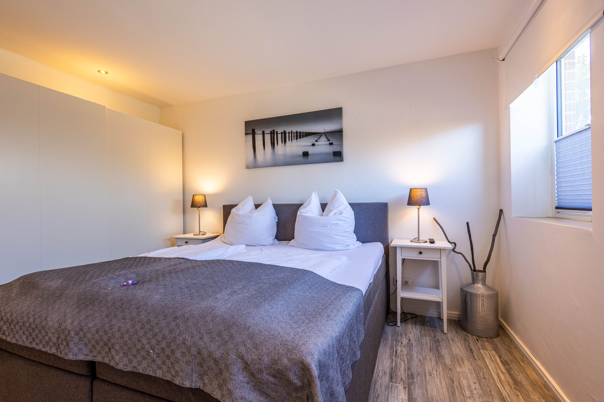 Schlafzimmer mit Doppelbett, grauer Tagesdecke, zwei Nachttischlampen und Wandbild mit Seeblick-Motiv