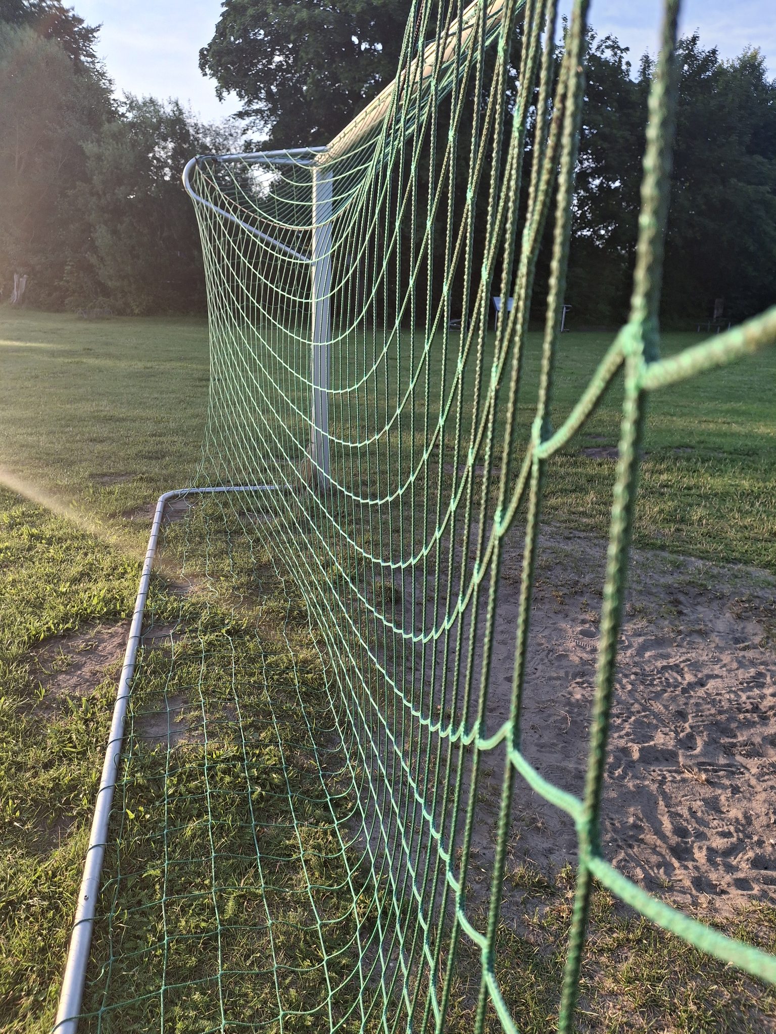 Nahaufnahme eines grünen Fußballtornetzes im Abendlicht auf einer Wiese, im Hintergrund ein zweites Tor