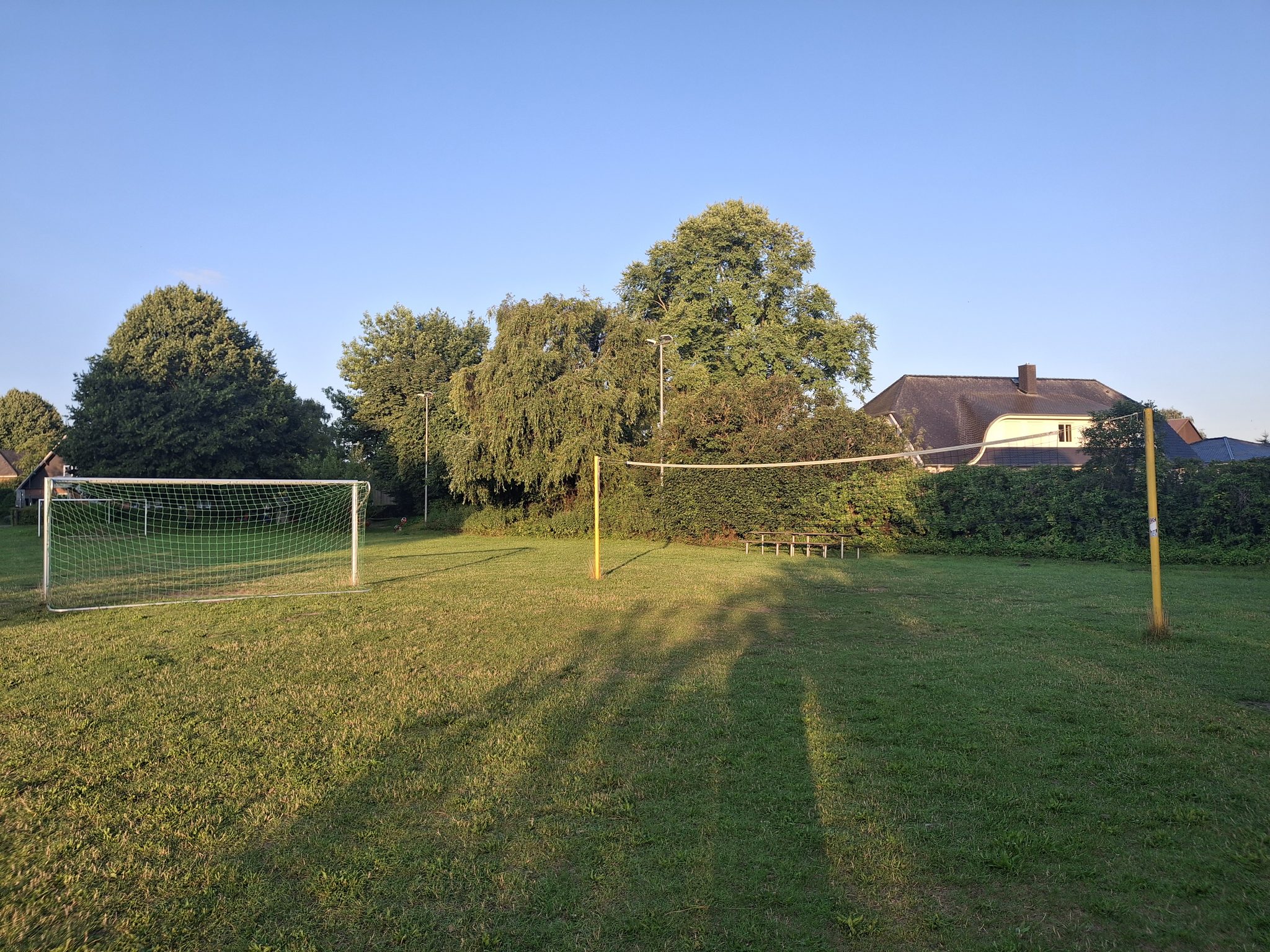 Grüne Wiese mit Fußballtor und Volleyballnetz im Abendlicht, umgeben von Bäumen und Hecken