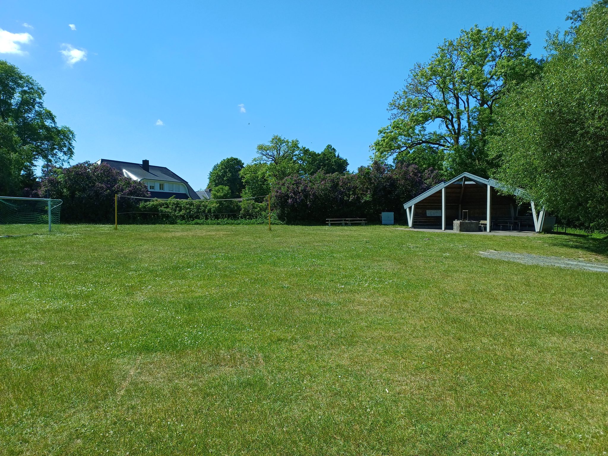 Große Rasenfläche mit Fußballtor, Volleyballnetz und überdachtem Pavillon, umgeben von Bäumen bei blauem Himmel