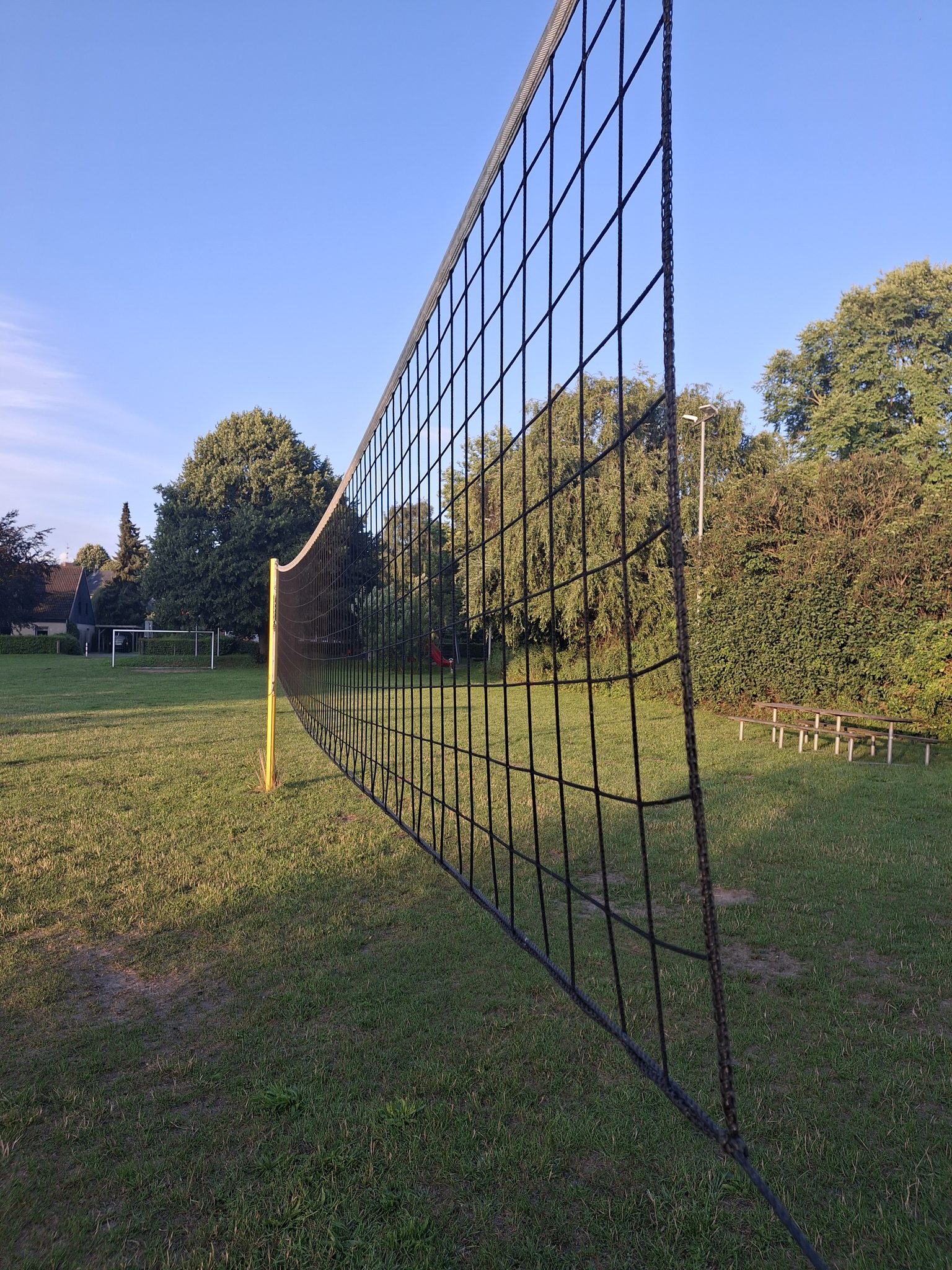 Volleyballnetz auf einer Grünfläche mit gelben Pfosten, im Hintergrund Bäume und blauer Himmel