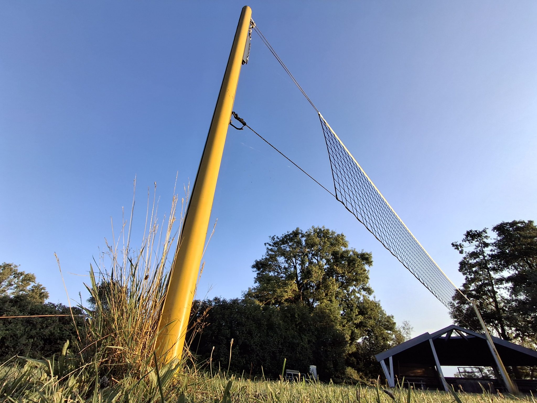 Gelber Volleyballpfosten mit gespanntem Netz auf der Seewiese, blauer Himmel und Bäume im Hintergrund