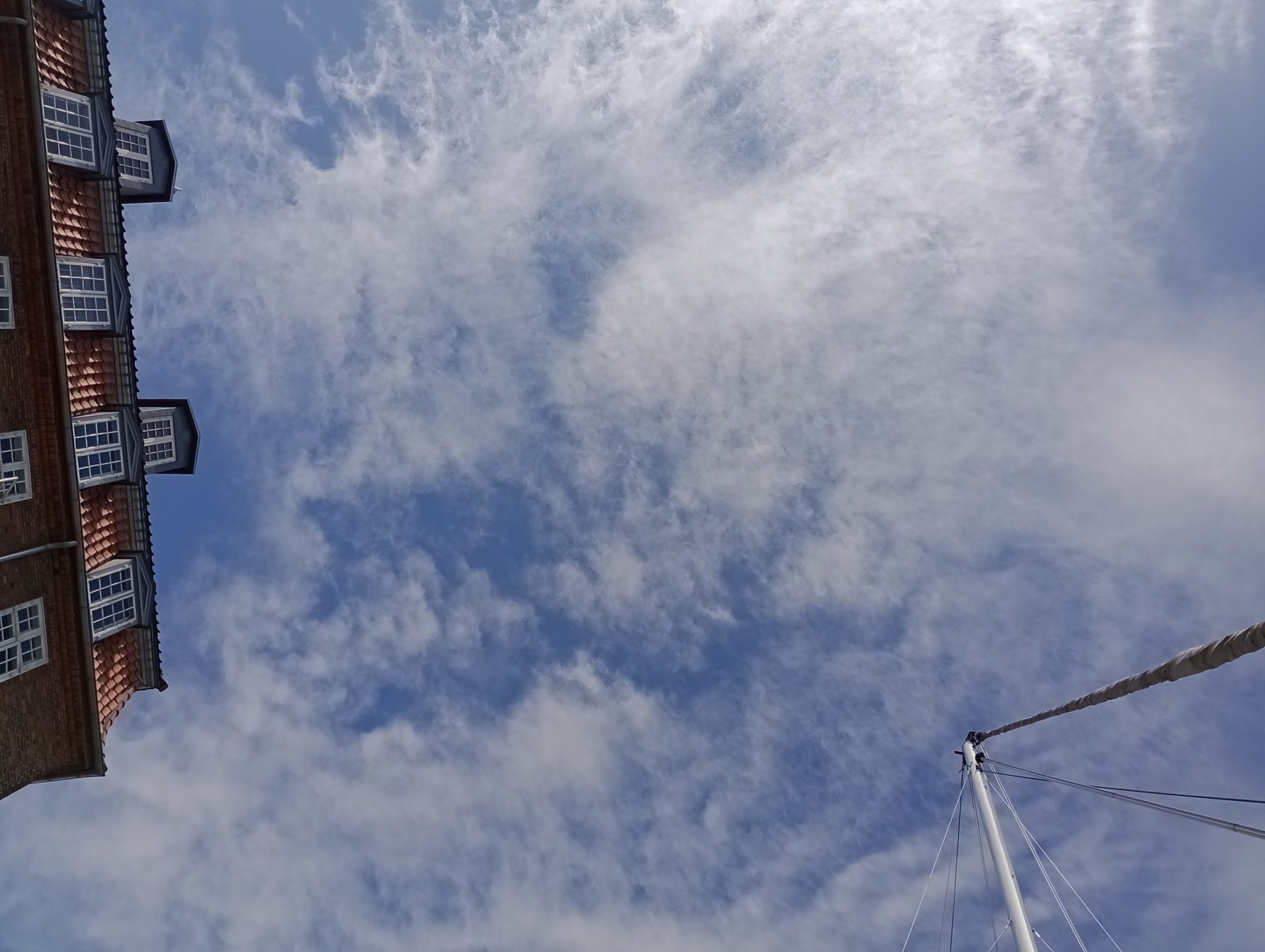 Froschperspektive: Backsteingebäude mit Gauben links, Segelmast rechts, blauer Himmel mit Schleierwolken