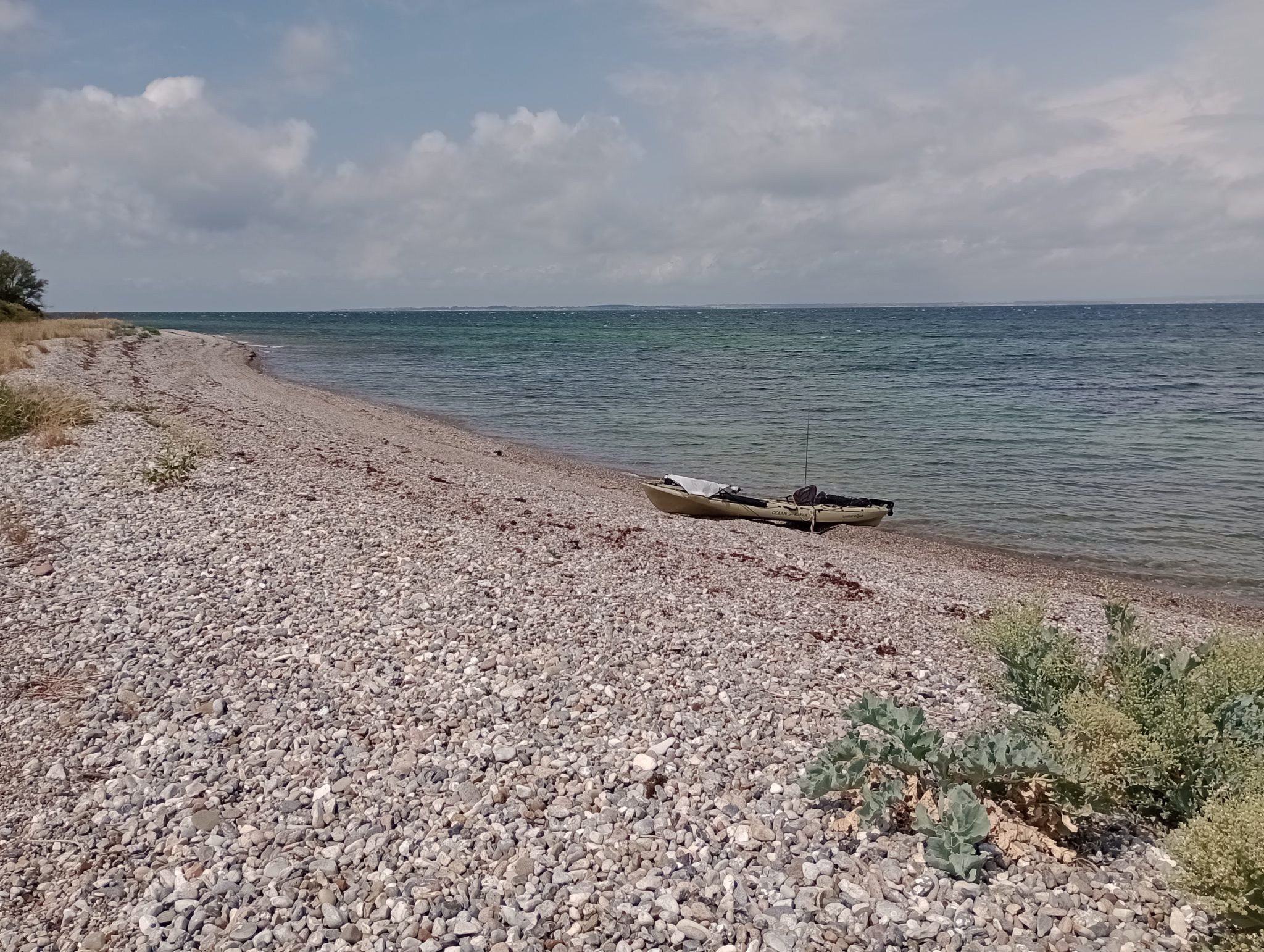 Kiesstrand mit einem Kajak am Wasser unter bewölktem Himmel, weite Ostseeküste