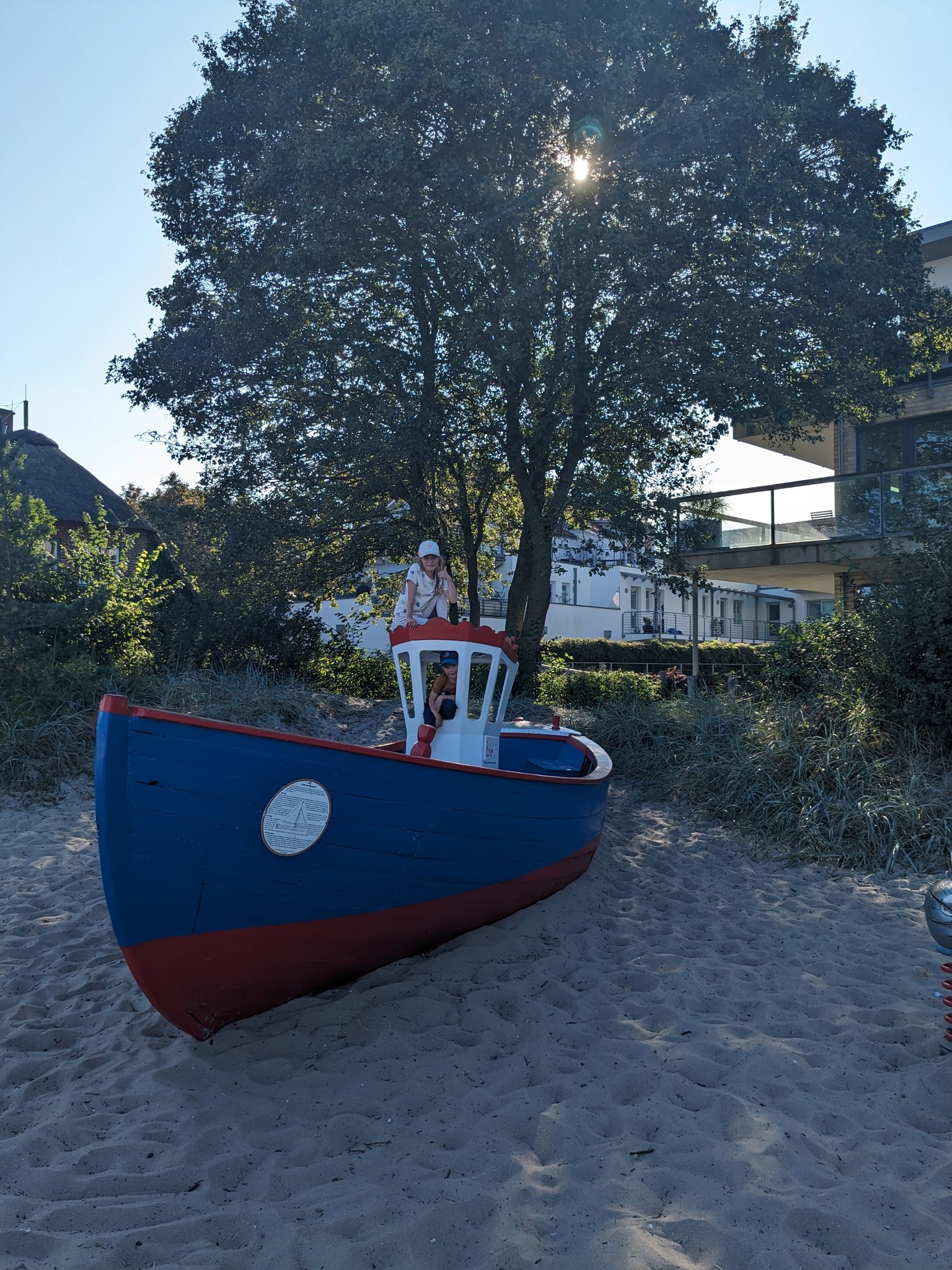 Kinder spielen auf einem blau-roten Spielboot im Sand, umgeben von Bäumen und Gebäuden im Hintergrund.