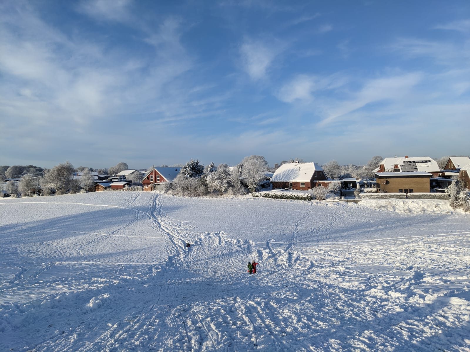 Verschneite Wiese mit Schlittenspuren und Kindern, dahinter schneebedeckte Häuser und Bäume unter blauem Winterhimmel
