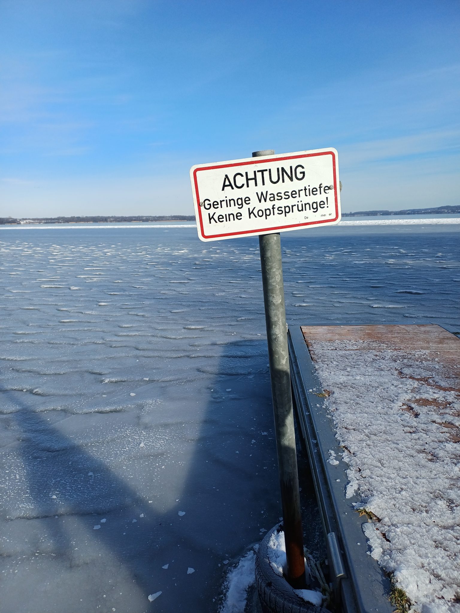 Warnschild 'Achtung – Geringe Wassertiefe, Keine Kopfsprünge' an einem Steg am zugefrorenen Wittensee im Winter
