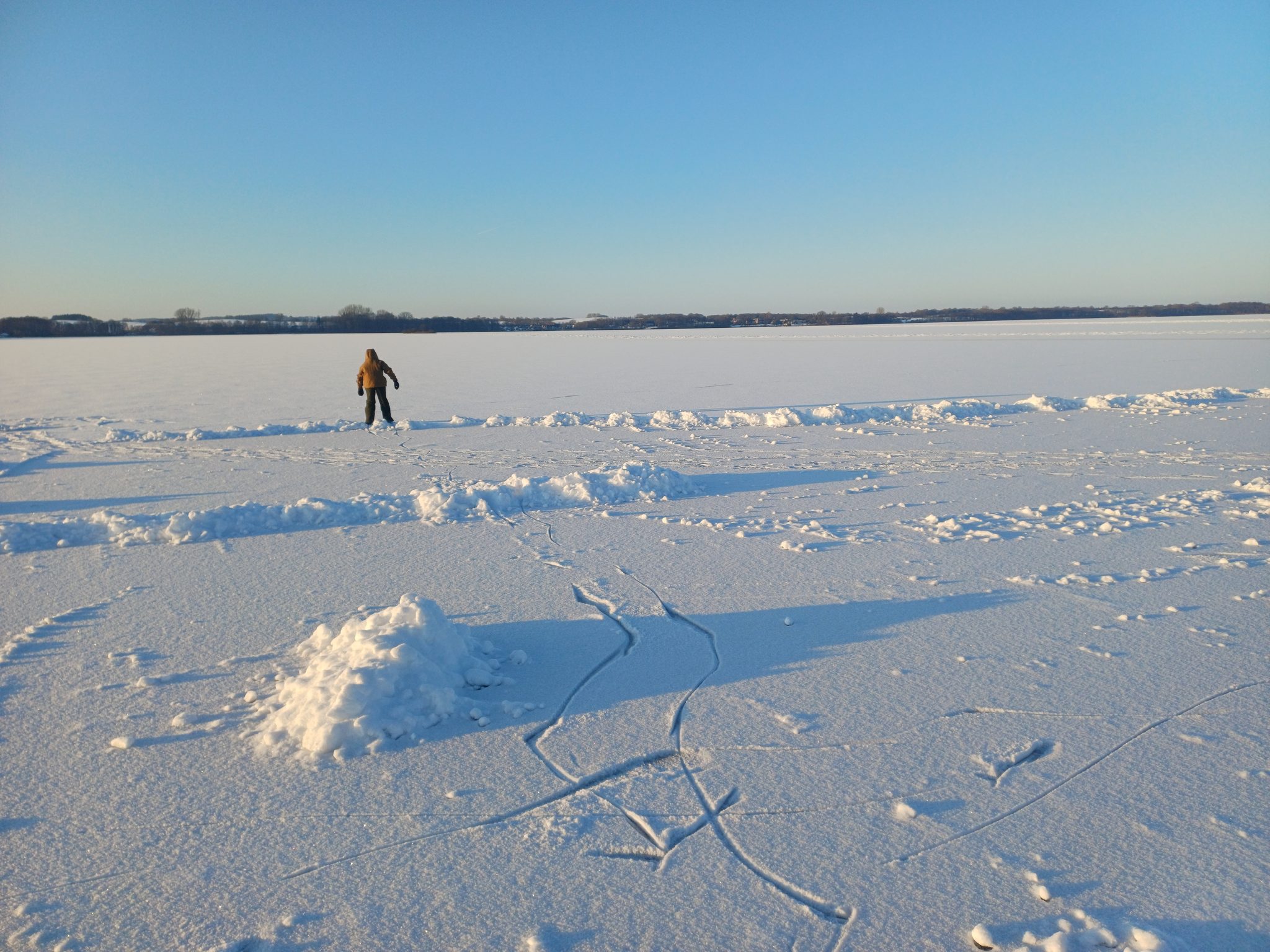Person mit gelber Jacke läuft auf dem schneebedeckten, zugefrorenen Wittensee bei blauem Winterhimmel