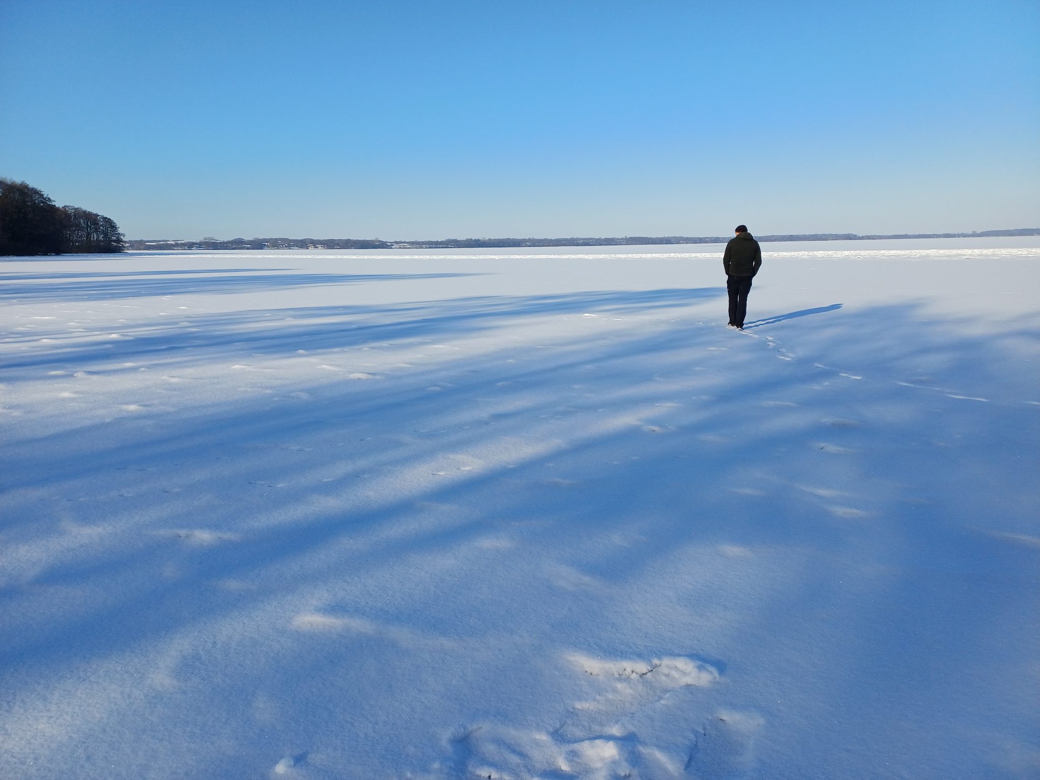 Person spaziert auf dem zugefrorenen, schneebedeckten Wittensee bei blauem Winterhimmel