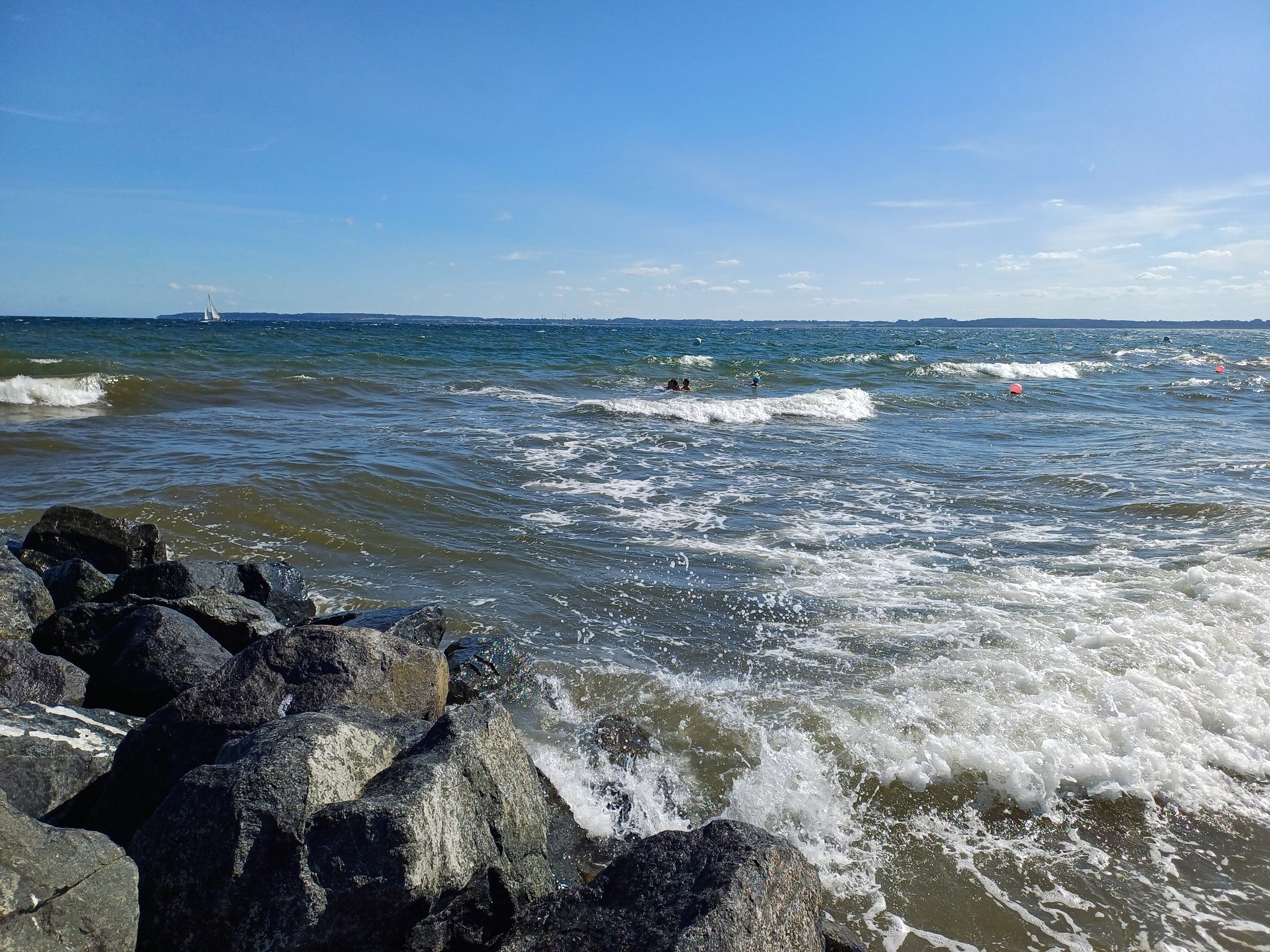Wellen brechen an Felsen am Wittensee, Schwimmer und Segelboot bei blauem Himmel